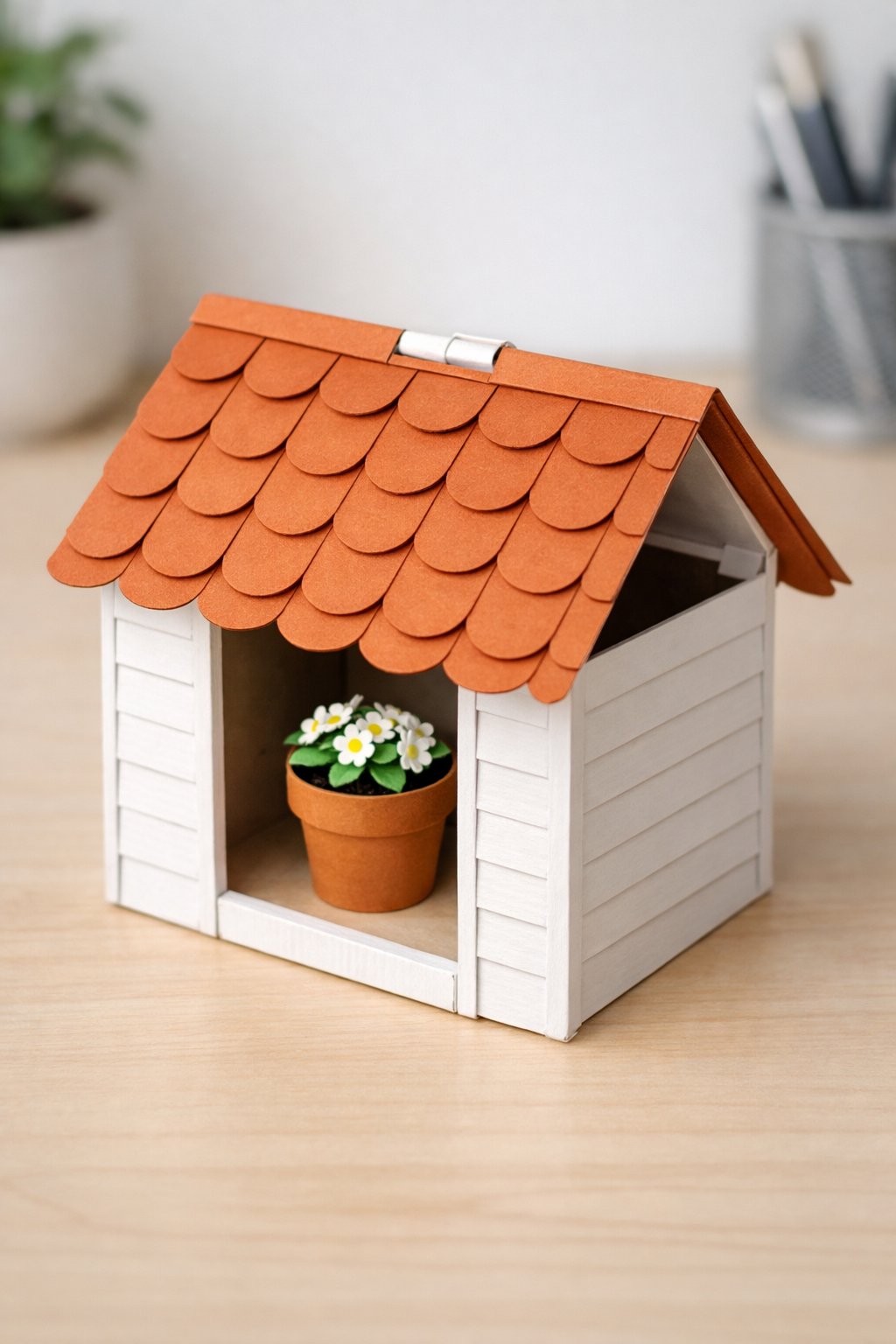 A 3D paper structure shaped like a small garden shed or birdhouse, standing on a desk. The roof is made of scalloped shingles in terracotta paper. The walls are white "shiplap" texture (horizontal lines). The roof is hinged and is shown slightly open, revealing a hollow interior where a small paper flower pot sits. The card is viewed from a slightly high angle to show the roof mechanism. No text or numbers on the house.