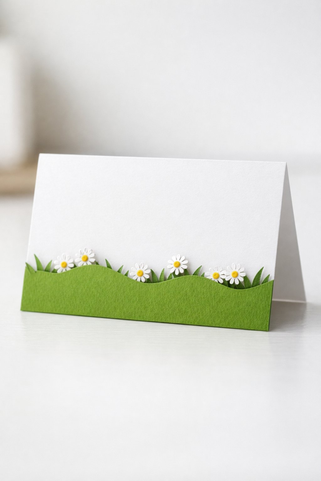 A realistic, bright photo of a horizontal "tent" style card. The bottom half of the card front is die-cut into a wavy grass shape with tiny paper daisies "peeking" over the edge. It stands independently on a tabletop, looking clean and modern.