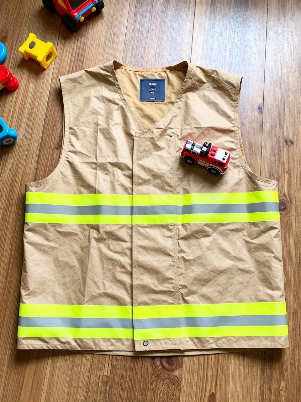 Photo of a paper bag firefighter vest laid flat on the floor, overhead flat lay, setting: a hardwood floor with scattered toys, lighting: natural morning light, containing one lived-in detail: a small plastic fire truck parked on the vest, iPhone photo quality.
