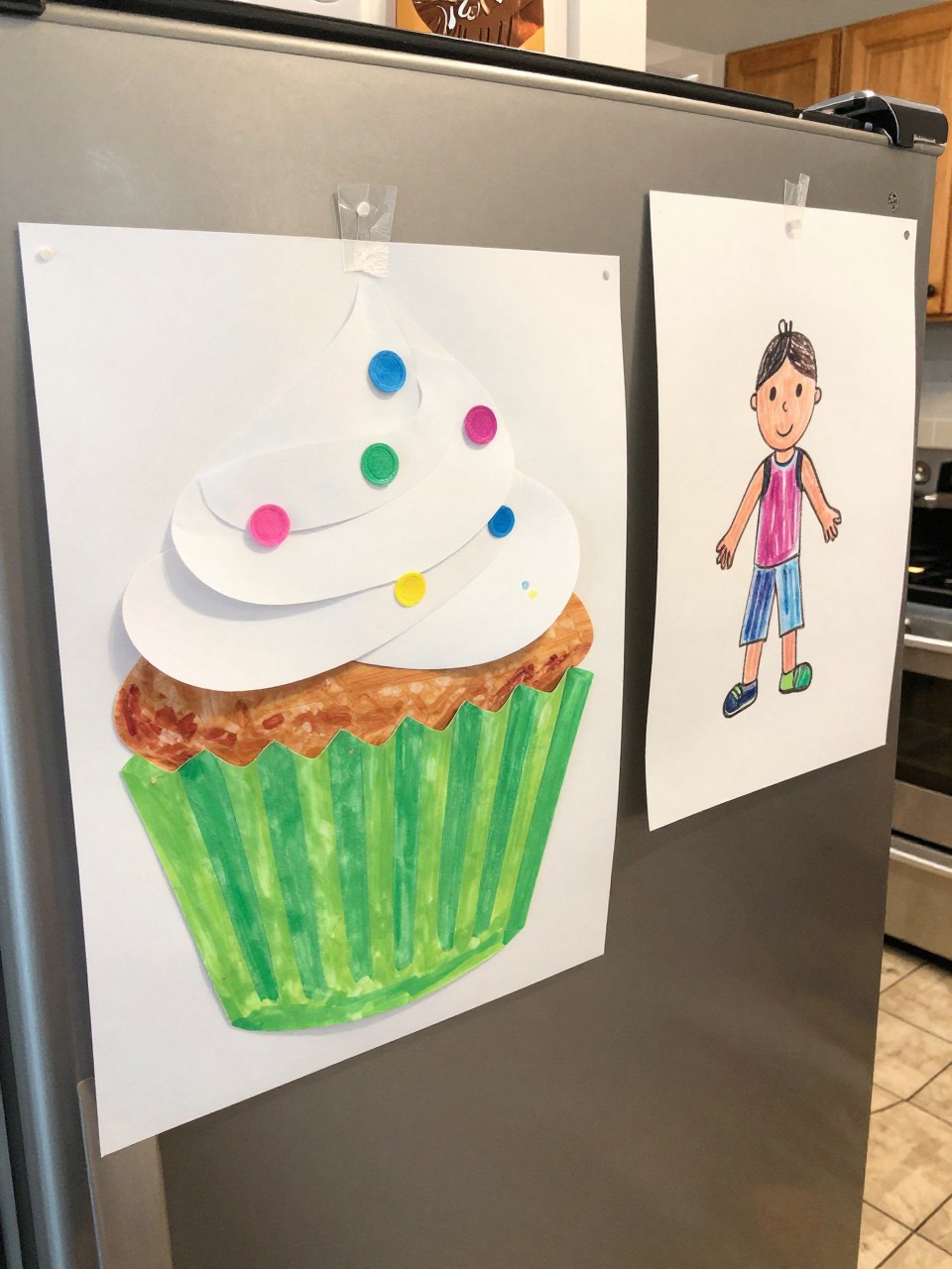 Photo of a cupcake liner teacher craft on a refrigerator, eye-level close-up, setting: a kitchen with a tile floor, lighting: bright kitchen light, containing one lived-in detail: a child's drawing hanging right next to it, iPhone photo quality.