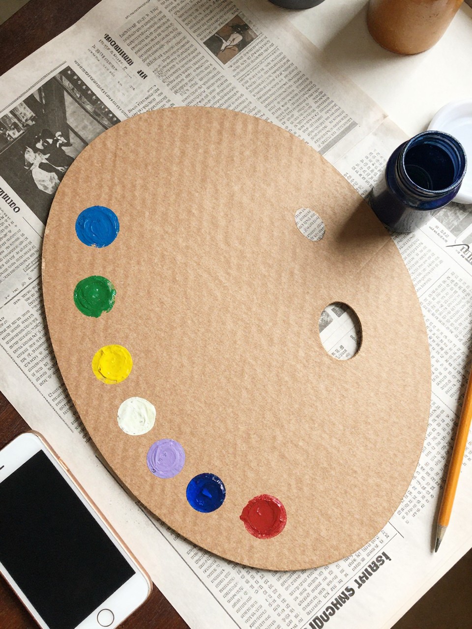 Photo of a cardboard artist palette craft with paint dots, overhead flat lay, setting: a messy art table with newspaper, lighting: natural morning light, containing one lived-in detail: an open paint bottle with a dried drip on the side, iPhone photo quality.