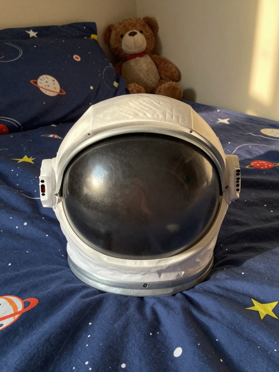Photo of a paper plate astronaut helmet resting on a bed, eye-level close-up, setting: a child's bedroom with space-themed sheets, lighting: warm afternoon light, containing one lived-in detail: a stuffed animal propped against the pillow, iPhone photo quality.