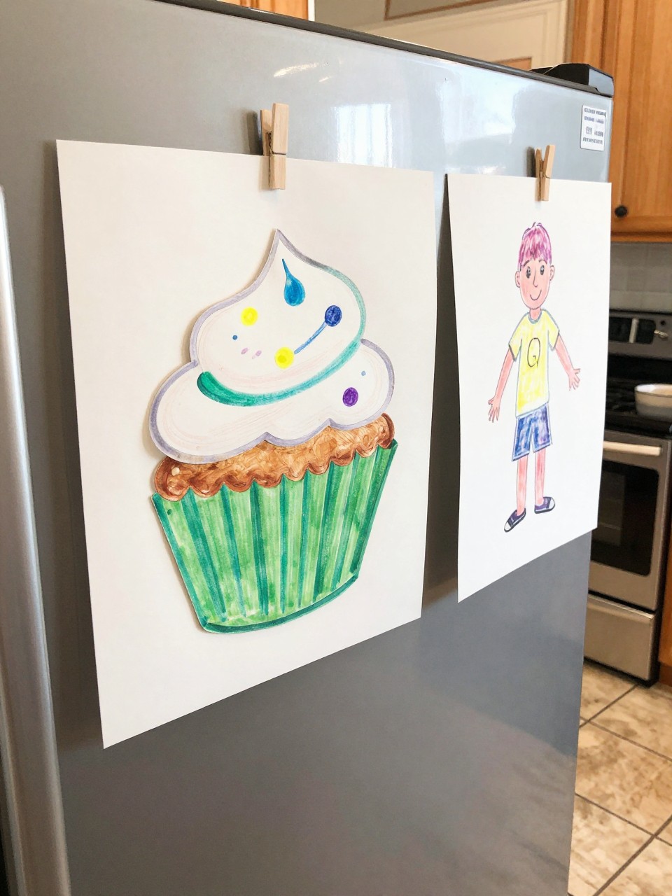 Photo of a cupcake liner teacher craft on a refrigerator, eye-level close-up, setting: a kitchen with a tile floor, lighting: bright kitchen light, containing one lived-in detail: a child's drawing hanging right next to it, iPhone photo quality.
