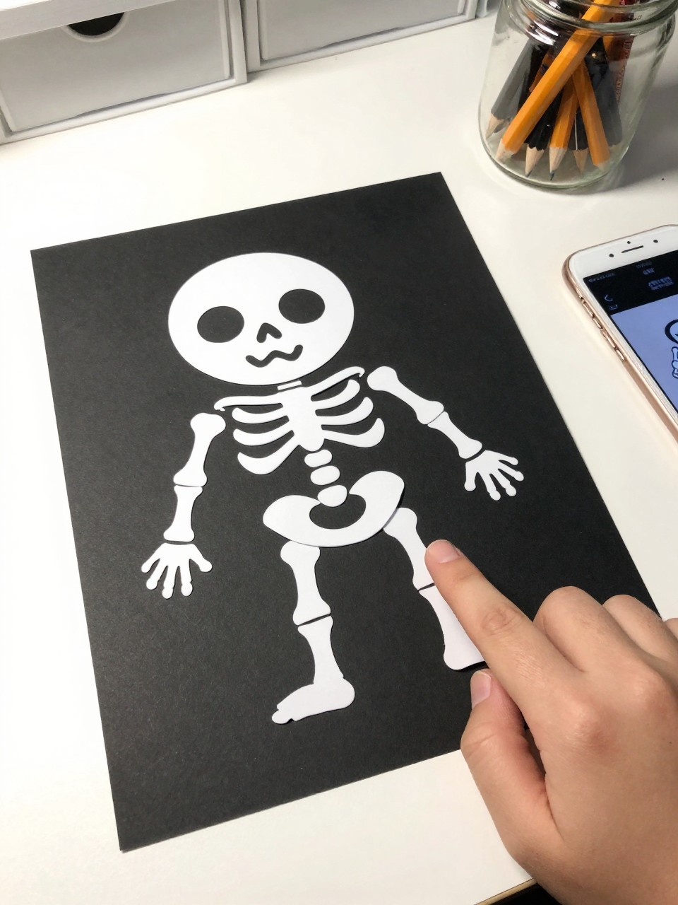Photo of a Q-tip skeleton craft on black paper, eye-level close-up, setting: a child's desk with a jar of pencils, lighting: bright overhead light, containing one lived-in detail: a small hand reaching into the frame to point at it, iPhone photo quality.