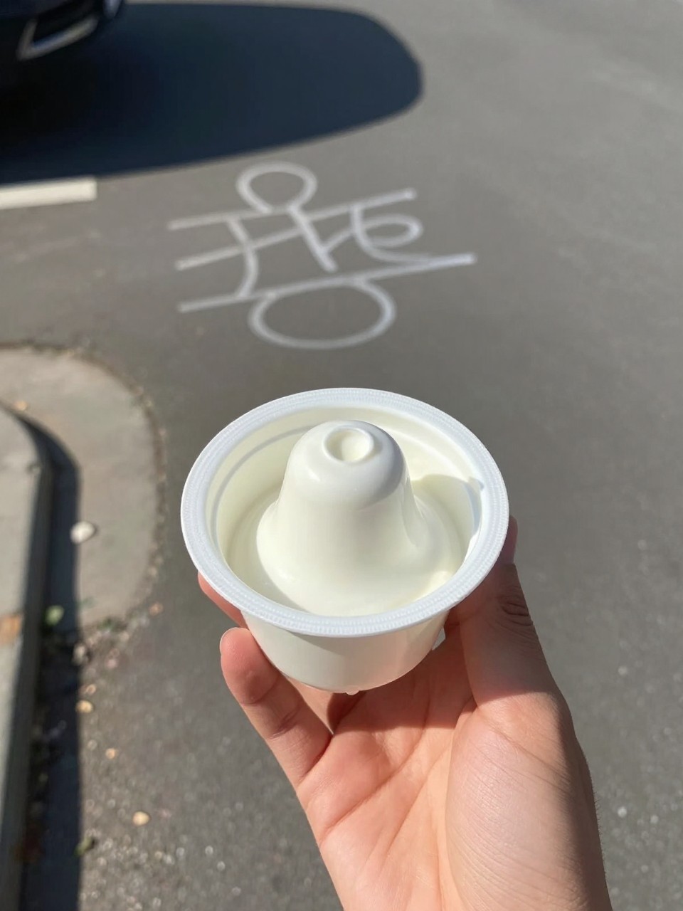 Photo of a finished yogurt cup bell being held in the air, held in hand (POV), setting: a driveway or sidewalk outside, lighting: bright afternoon sun, containing one lived-in detail: a chalk drawing on the ground in the background, iPhone photo quality.