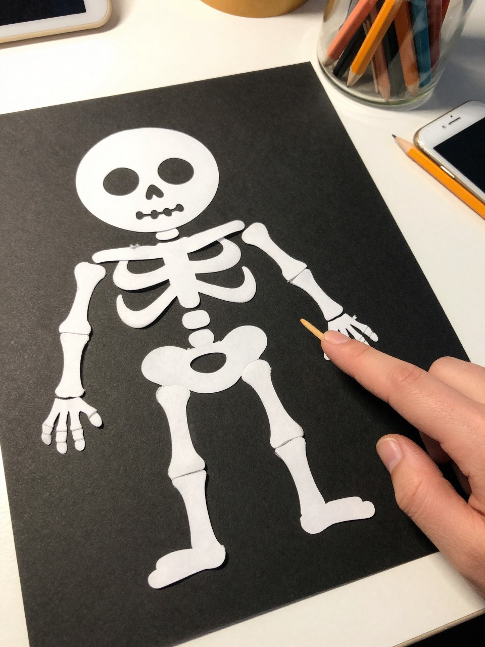 Photo of a Q-tip skeleton craft on black paper, eye-level close-up, setting: a child's desk with a jar of pencils, lighting: bright overhead light, containing one lived-in detail: a small hand reaching into the frame to point at it, iPhone photo quality.
