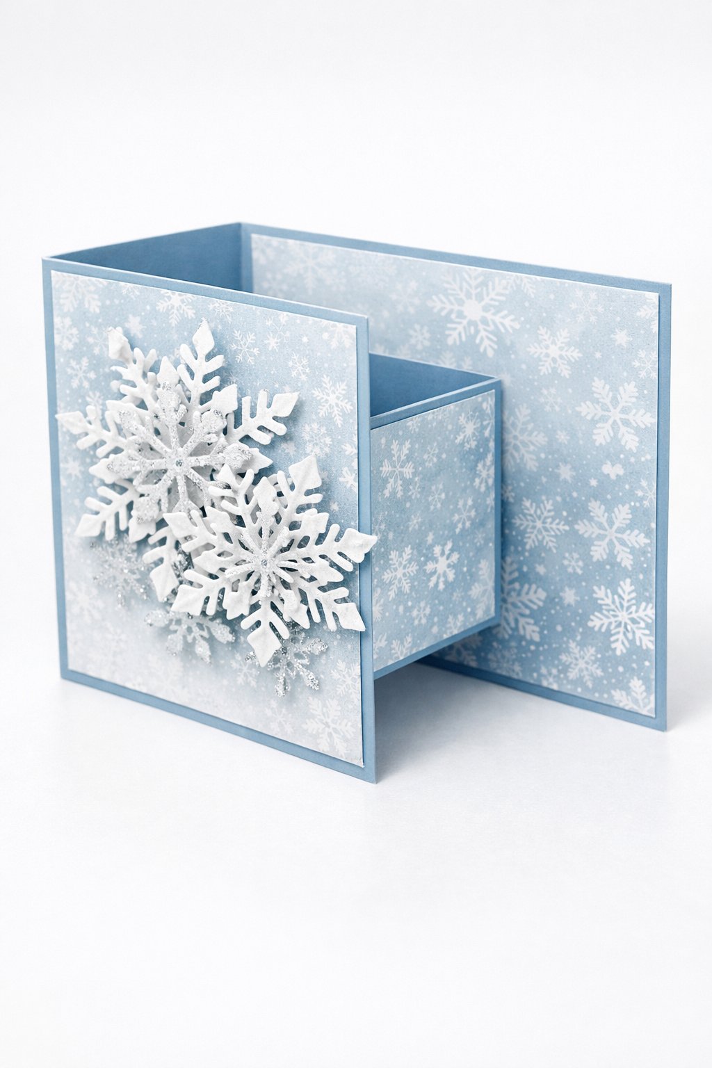 Ultra realistic product photo of a handmade double Z fold card, shown partially open to display the fold structure, icy blue and white color palette, snowflake patterned paper on panels, layered white snowflake die-cuts on the front panel with subtle silver shimmer accents (no lettering), crisp folds, matte cardstock texture, photographed on a bright white surface with cool soft lighting, minimal props, no hands, no text, no watermark, sharp focus, high detail.