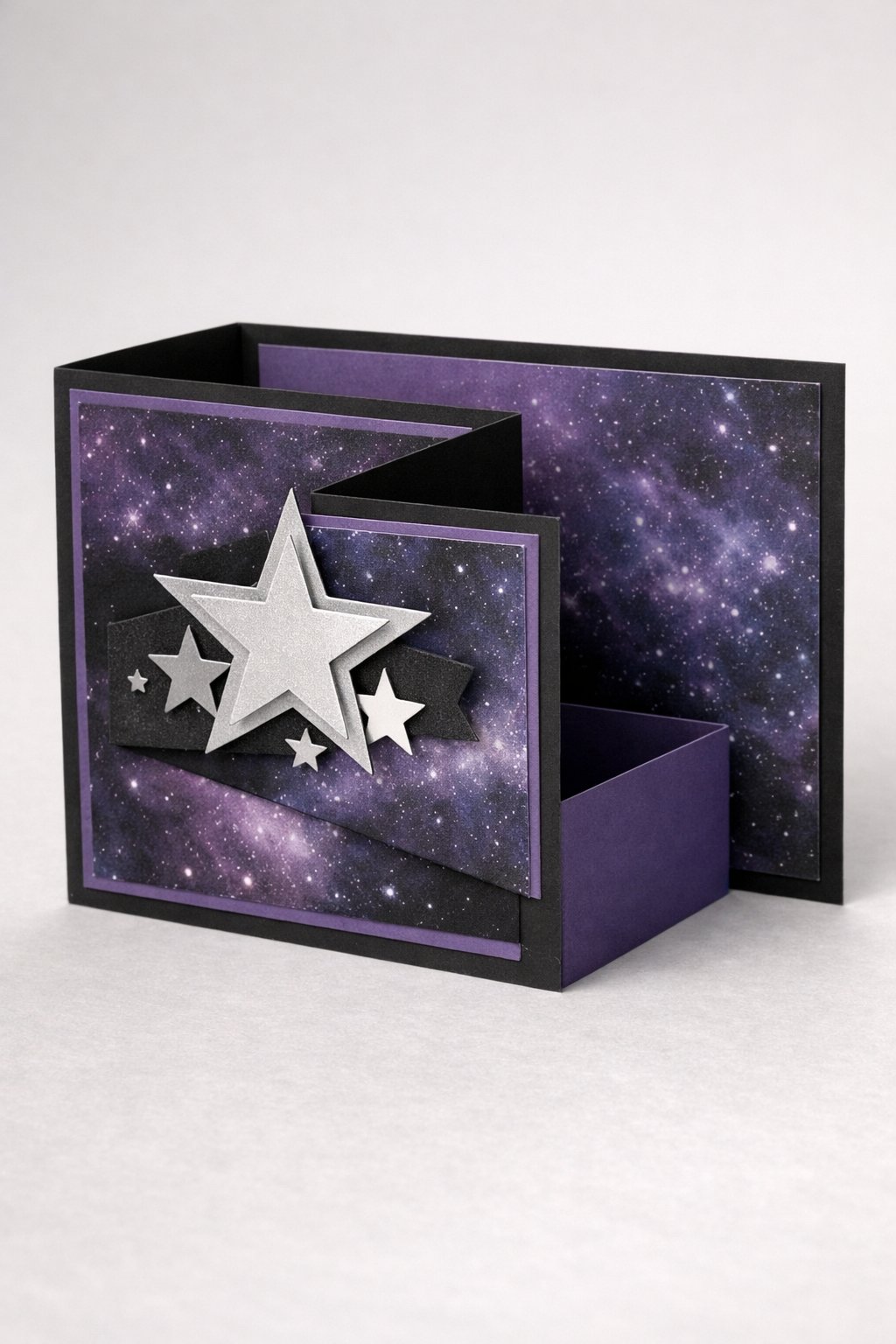 Ultra realistic product photo of a handmade double Z fold card, shown slightly open to clearly display the fold structure, deep purple and black color palette, galaxy patterned paper on panels with subtle star speckles, layered silver star cutouts on the front panel, crisp folds, matte cardstock texture, photographed on a clean neutral background with soft studio lighting, no hands, no text, no watermark, sharp focus, high detail.