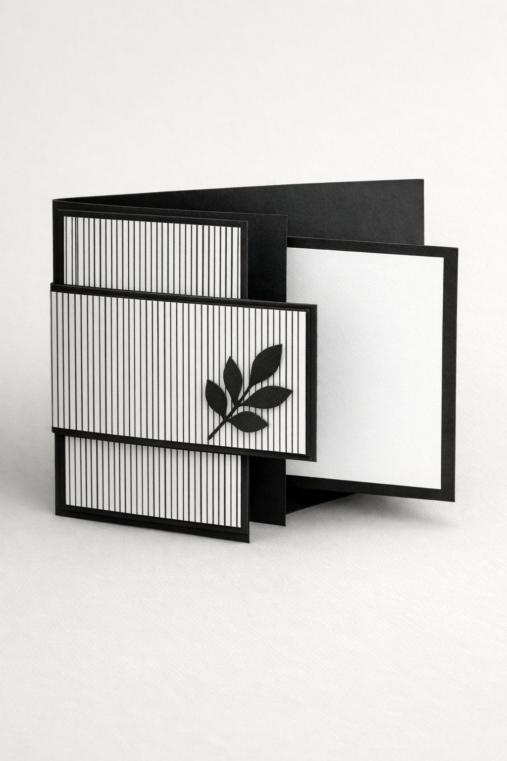 Ultra realistic product photo of a handmade joy fold card, shown slightly open to clearly display the wrap design and smaller inner card, clean black and white color palette, thin striped patterned paper on the wrap front, simple solid black mat layers, small single paper leaf sprig embellishment on the wrap flap, crisp folds, matte cardstock texture, photographed on a bright neutral background with soft studio lighting, no hands, no envelope, no text, no watermark, sharp focus, high detail.