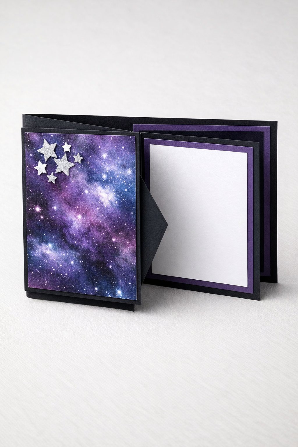 Ultra realistic product photo of a handmade joy fold card, shown slightly open to clearly display the wrap and the smaller inner card, deep navy and purple galaxy theme, galaxy patterned paper on the wrap front, layered dark cardstock mats, small silver star paper cutouts clustered on the wrap flap, crisp folds, matte cardstock texture, photographed on a clean light neutral background with soft studio lighting, no hands, no text, no watermark, sharp focus, high detail.