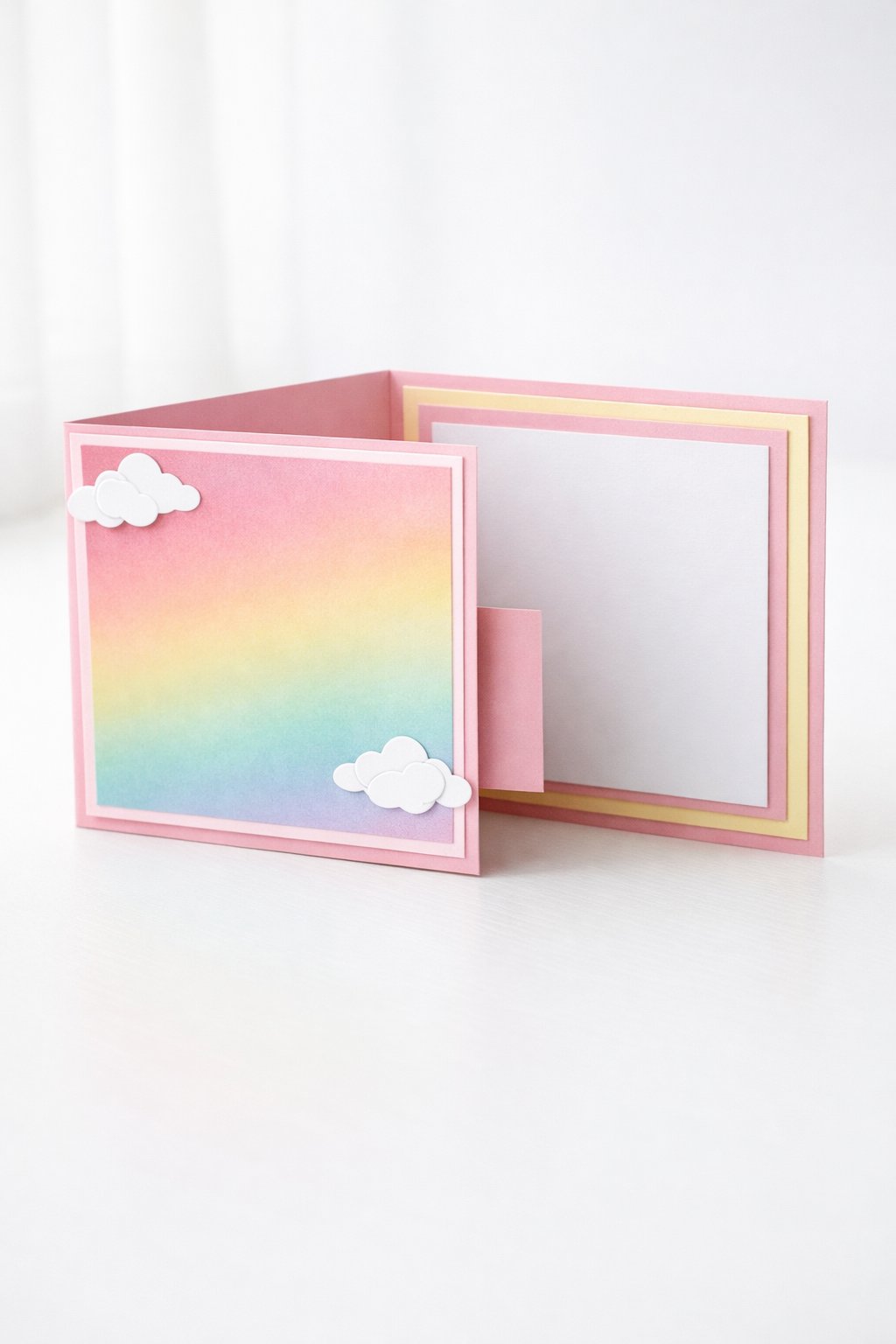 Ultra realistic product photo of a handmade joy fold card, shown slightly open to clearly display the wrap and inner card, pastel rainbow theme with soft gradient paper on the wrap front, small white cloud paper cutouts on the wrap flap corner, layered pastel mats, crisp folds, matte cardstock texture, photographed on a bright white tabletop with soft natural window light, minimal props, no hands, no text, no watermark, sharp focus, high detail.