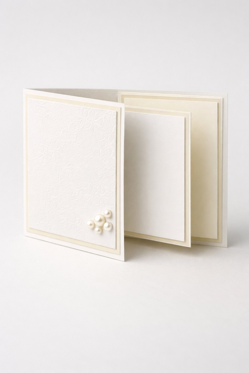 Ultra realistic product photo of a handmade joy fold card, shown slightly open to clearly display the wrap-around outer panel and the smaller inner card, pure white and soft ivory color palette, subtle embossed texture on the wrap front panel, thin ivory mat layers, small cluster of pearl flat-back half beads on the wrap flap corner, crisp folds, matte cardstock texture, photographed on a clean light background with soft studio lighting, minimal props, no hands, no envelope, no text, no watermark, sharp focus, high detail.
