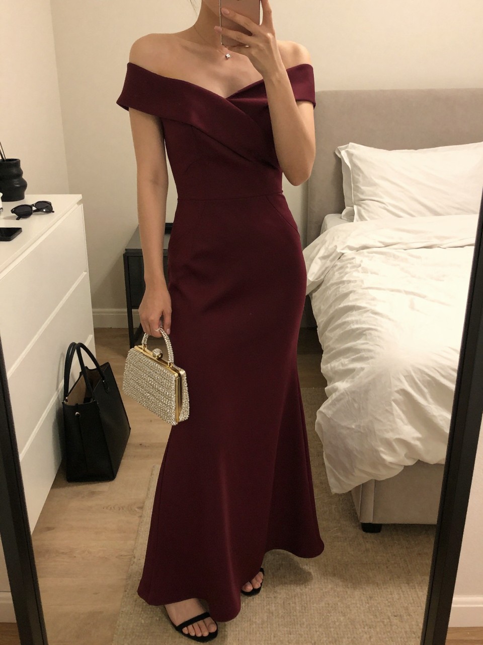 mirror selfie of a woman wearing a burgundy off-shoulder floor-length gown in satin or crepe, black strappy heels, small crystal stud earrings, and a sparkly beaded clutch, holding a phone in front of a mirror, cropped at NECK so face doesn't show, with a natural casual selfie pose, in a feminine-themed bedroom, warm moody lighting, including small lived-in details like a tote bag on the floor, sunglasses on a dresser, or soft bedding in the background, latest iPhone photo quality.