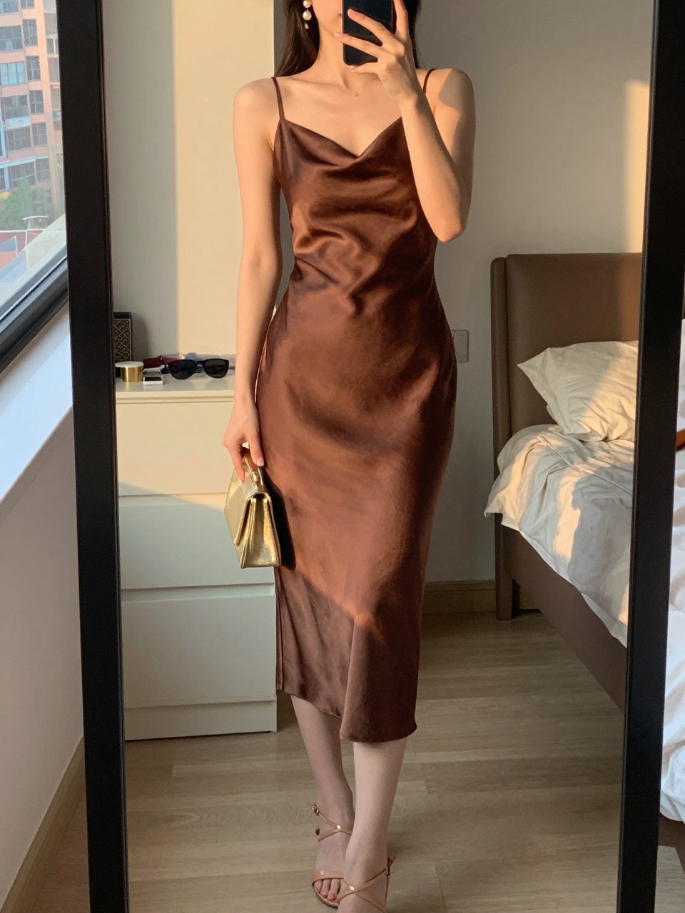 mirror selfie of a woman wearing a chocolate brown satin slip midi dress with a bias cut, pearl drop earrings, nude strappy heels, and a small gold clutch, holding a phone in front of a mirror, cropped at NECK so face doesn't show, with a natural casual selfie pose, in a city apartment, evening golden hour, including small lived-in details sunglasses on a dresser, or soft bedding in the background, latest iPhone photo quality.