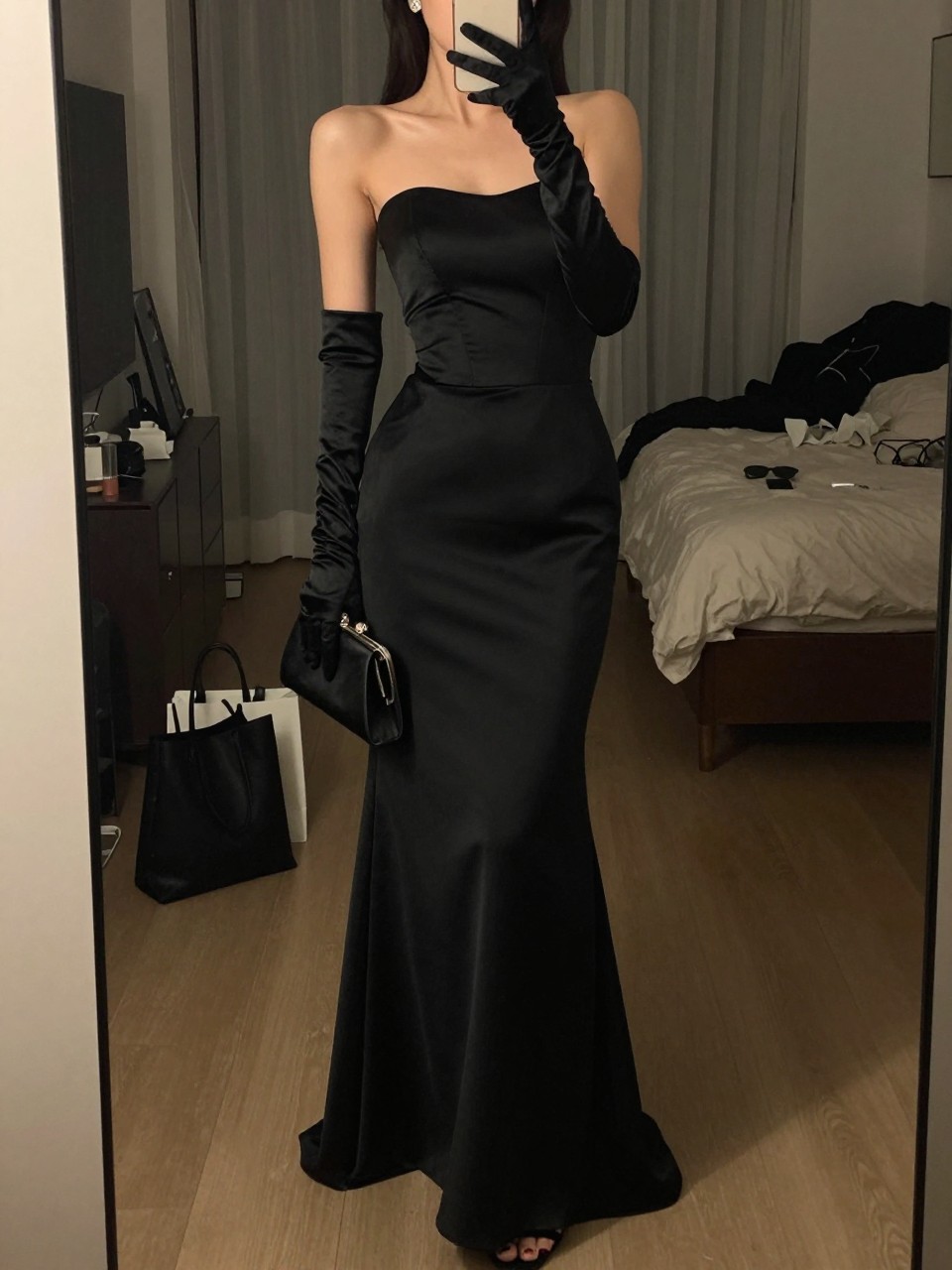 mirror selfie of a woman wearing a black strapless structured satin floor-length gown, black satin opera gloves, black strappy stilettos, simple diamond stud earrings, and a black satin clutch, holding a phone in front of a mirror, cropped at NECK so face doesn't show, with a natural casual selfie pose, in a city apartment, warm moody lighting, including small lived-in details like a tote bag on the floor, sunglasses on a dresser, or soft bedding in the background, latest iPhone photo quality.