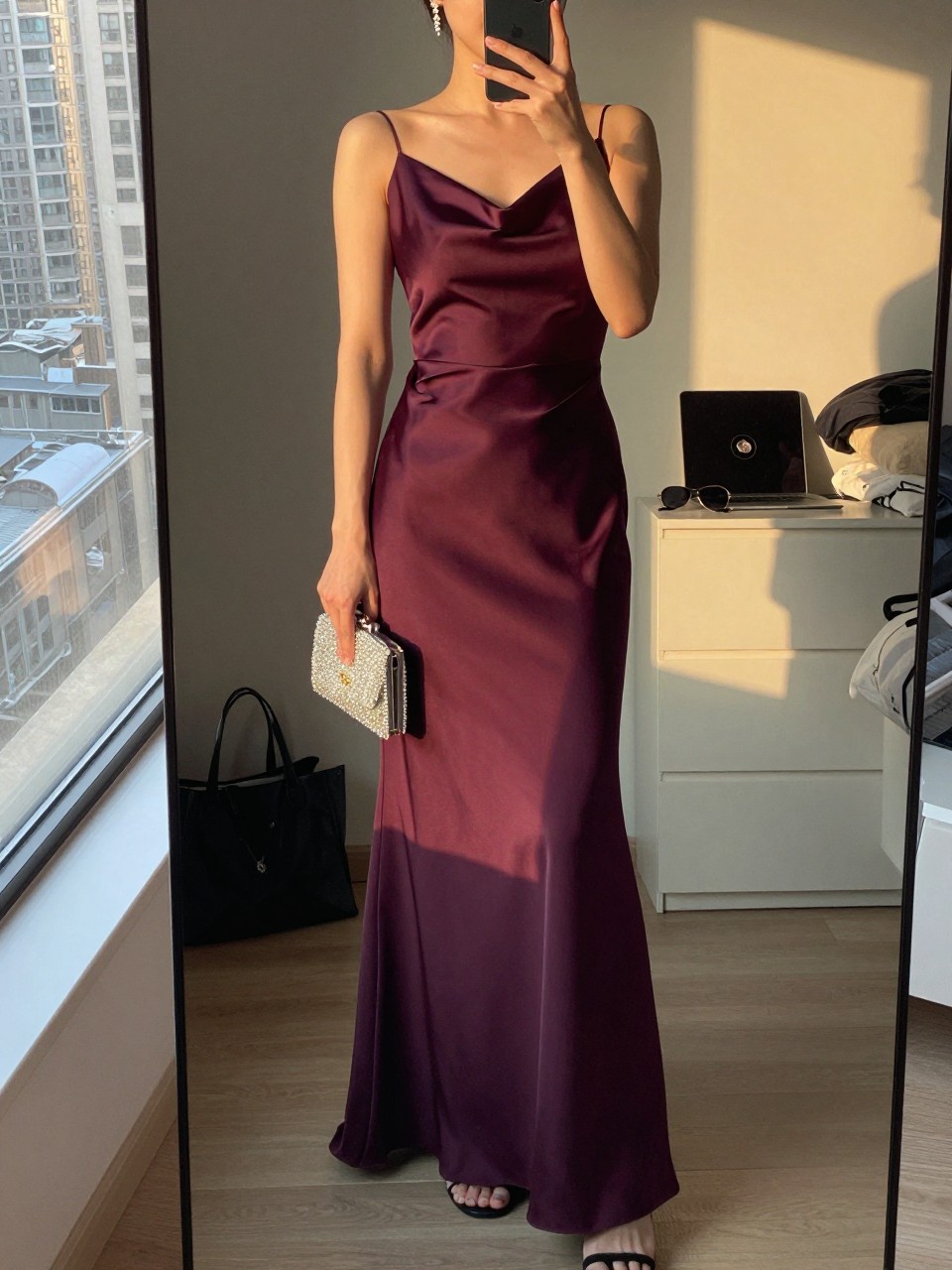 mirror selfie of a woman wearing a deep plum floor-length satin gown with a slight drape, black strappy heels, small chandelier earrings, and a beaded evening clutch, holding a phone in front of a mirror, cropped at NECK so face doesn't show, with a natural casual selfie pose, in a city apartment, evening golden hour, including small lived-in details like a tote bag on the floor, sunglasses on a dresser, or soft bedding in the background, latest iPhone photo quality.