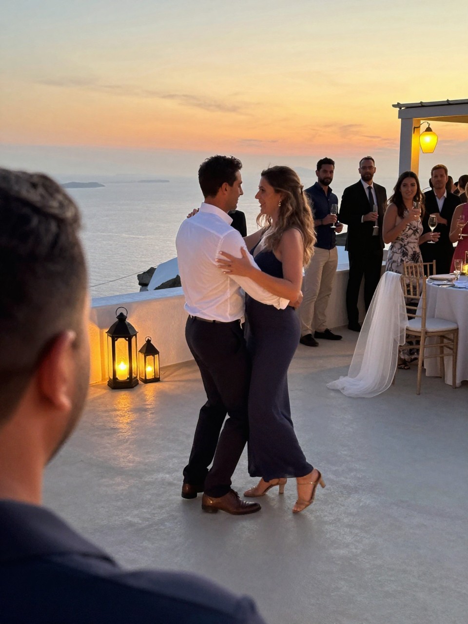 Photo of a couple slow dancing on a Mediterranean terrace at sunset with a few lanterns nearby and guests watching softly in the background, camera angle: over-the-shoulder candid, setting: terrace dance area, lighting: golden hour, containing subtle real wedding activity such as guests holding champagne flutes and a veil draped over a nearby chair, latest iPhone photo quality.