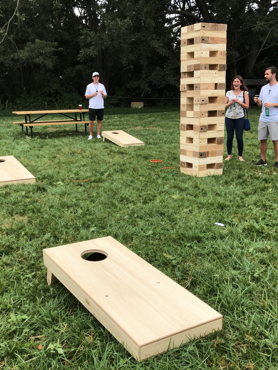 Photo of an outdoor camp field set up with cornhole boards, giant Jenga, and ring toss with picnic tables nearby, camera angle: wide ceremony view, setting: camp games field, lighting: natural daylight, containing subtle real wedding activity such as guests playing cornhole and laughing while holding drinks, latest iPhone photo quality.