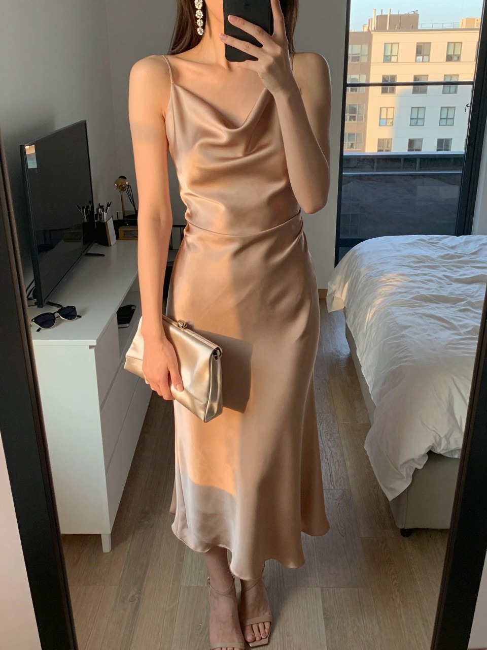 mirror selfie of a woman wearing a tan satin midi dress with a draped neckline, crystal drop earrings, nude strappy heels, and a champagne satin clutch, holding a phone in front of a mirror, cropped at NECK so face doesn't show, with a natural casual selfie pose, in a city apartment, evening golden hour, including small lived-in details like sunglasses on a dresser, or soft bedding in the background, latest iPhone photo quality.
