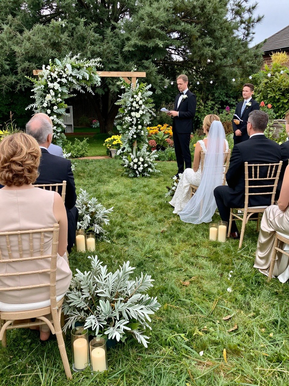 Photo of a late summer garden ceremony setup with sage greenery arrangements, champagne-toned chairs, and soft candle accents, camera angle: wide ceremony view, setting: outdoor garden ceremony aisle, lighting: natural daylight, containing subtle real wedding activity such as guests finding their seats and a veil draped over a chair, latest iPhone photo quality.