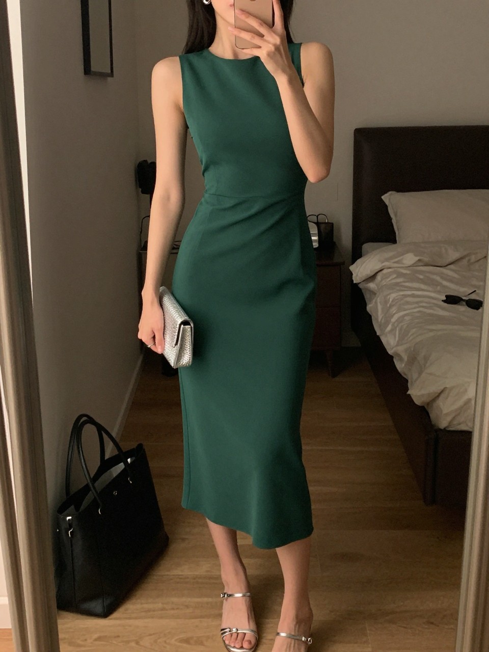 mirror selfie of a woman wearing a forest green crepe midi dress, silver strappy heels, silver stud earrings, and a small silver clutch, holding a phone in front of a mirror, cropped at NECK so face doesn't show, with a natural casual selfie pose, in a city apartment, warm moody lighting, including small lived-in details like a tote bag on the floor, sunglasses on a dresser, or soft bedding in the background, latest iPhone photo quality.