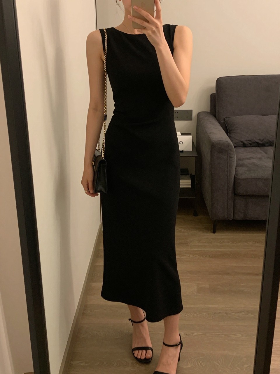 mirror selfie of a woman wearing a black lightweight crepe midi dress, black strappy heels, small gold stud earrings, and a gold chain clutch, holding a phone in front of a mirror, cropped at NECK so face doesn't show, with a natural casual selfie pose, in a city apartment, warm moody lighting, including small lived-in details.
