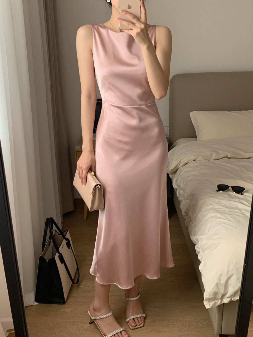mirror selfie of a woman wearing a soft pink satin midi dress with a clean neckline, crystal strap heels, simple crystal stud earrings, and a small blush clutch, holding a phone in front of a mirror, cropped at NECK so face doesn't show, with a natural casual selfie pose, in a feminine-themed bedroom, warm moody lighting, including small lived-in details like a tote bag on the floor, sunglasses on a dresser, or soft bedding in the background, latest iPhone photo quality.