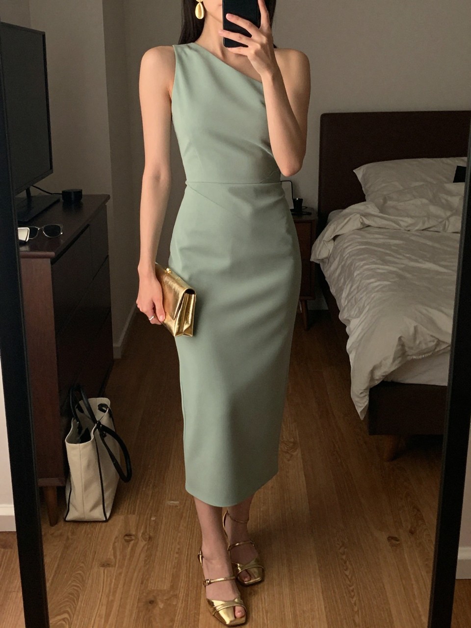 mirror selfie of a woman wearing a sage green crepe one-shoulder midi dress, gold statement drop earrings, metallic gold strappy heels, and a small gold clutch, holding a phone in front of a mirror, cropped at NECK so face doesn't show, with a natural casual selfie pose, in a city apartment, warm moody lighting, including small lived-in details like a tote bag on the floor, sunglasses on a dresser, or soft bedding in the background, latest iPhone photo quality.