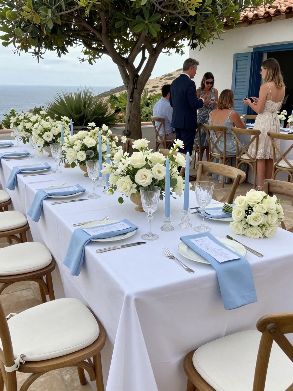 Photo of a late summer terrace reception table with soft white linens, sky blue napkins, white florals, and pale blue taper candles, camera angle: guest table angle, setting: Mediterranean terrace reception, lighting: natural daylight, containing subtle real wedding activity such as guests chatting and a bouquet resting on a nearby chair, latest iPhone photo quality.