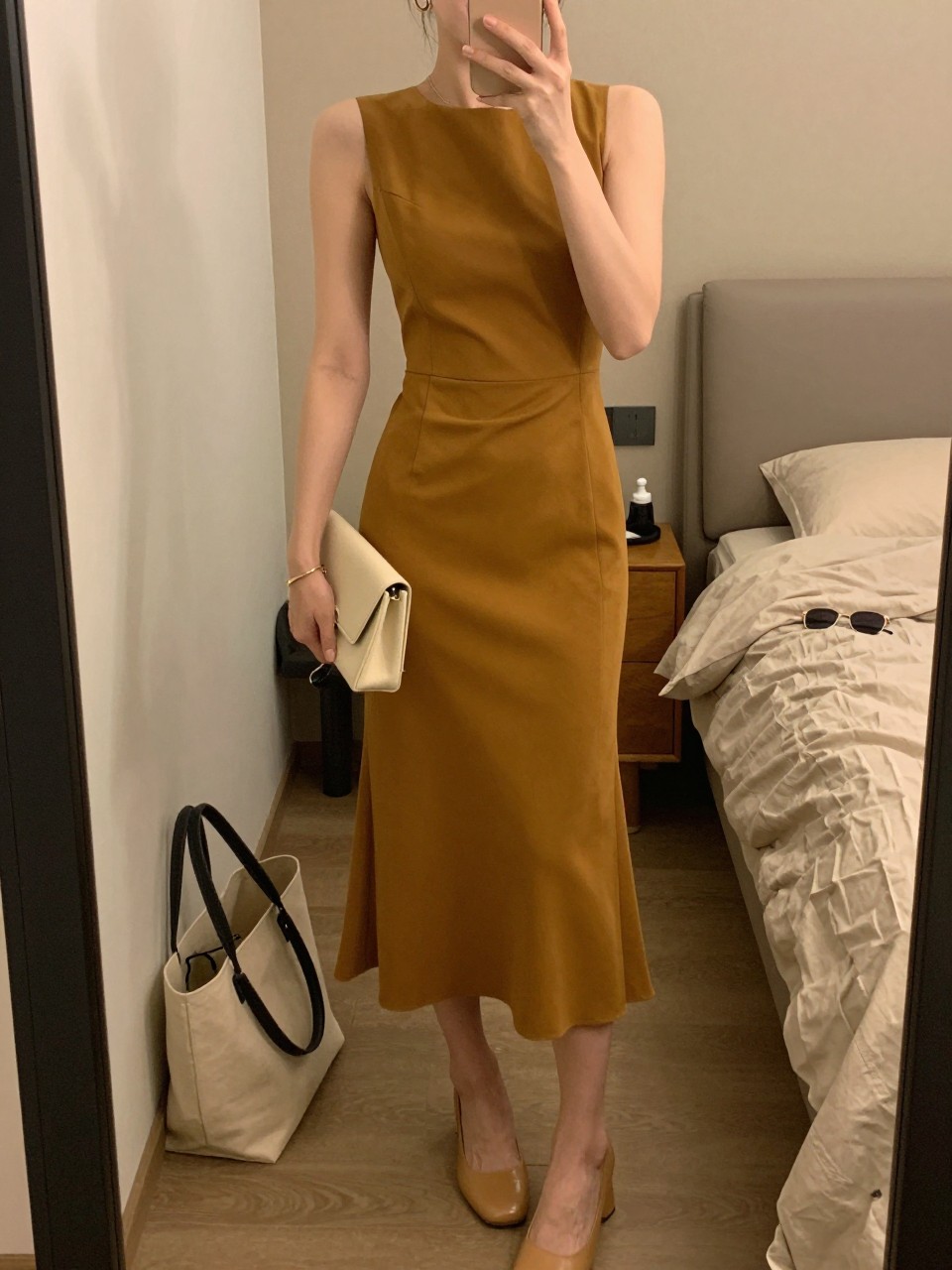 mirror selfie of a woman wearing a mustard crepe midi dress with a fitted waist, tan block heels, small gold hoop earrings, and a small cream clutch, holding a phone in front of a mirror, cropped at NECK so face doesn't show, with a natural casual selfie pose, in a feminine-themed bedroom, warm moody lighting, including small lived-in details like a tote bag on the floor, sunglasses on a dresser, or soft bedding in the background, latest iPhone photo quality.