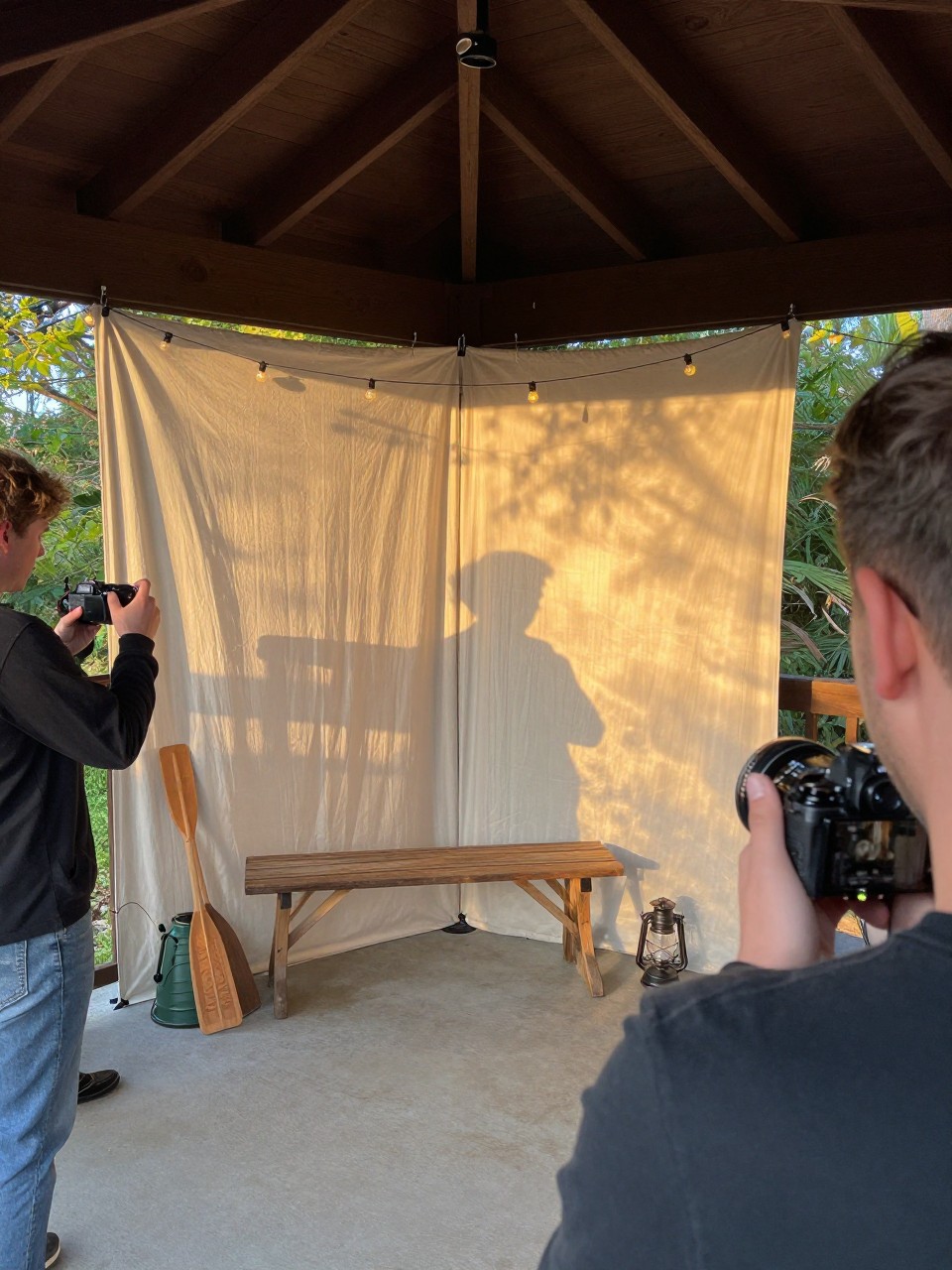 Photo of a cabin porch photo booth corner with a simple fabric backdrop, a bench, camp props like paddles and lanterns, and string lights above, camera angle: over-the-shoulder candid, setting: cabin porch area, lighting: golden hour, containing subtle real wedding activity such as guests taking turns posing and someone holding a camera nearby, latest iPhone photo quality.