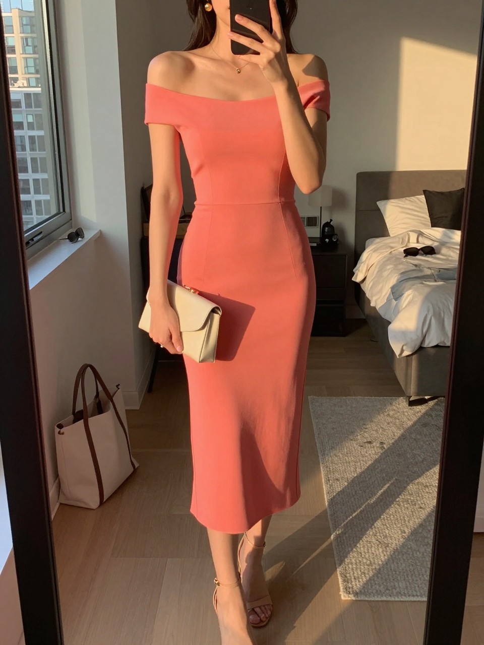 mirror selfie of a woman wearing a coral crepe off-shoulder midi dress, nude strappy heels, small gold drop earrings, and a small cream clutch, holding a phone in front of a mirror, cropped at NECK so face doesn't show, with a natural casual selfie pose, in a city apartment, evening golden hour, including small lived-in details like a tote bag on the floor, sunglasses on a dresser, or soft bedding in the background, latest iPhone photo quality.