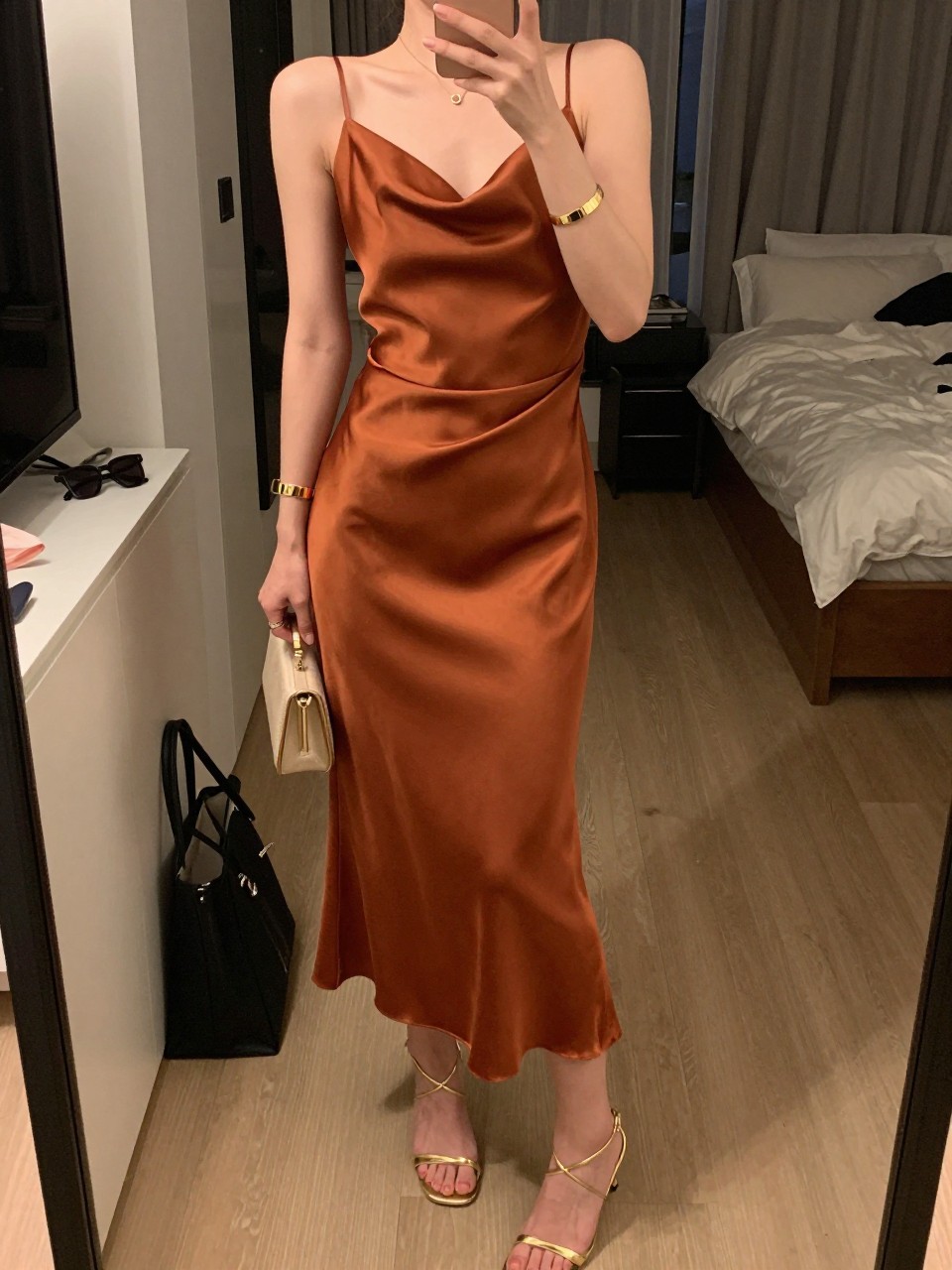 mirror selfie of a woman wearing a terracotta satin midi dress with a draped skirt, gold strappy heels, a slim gold cuff bracelet, and a small champagne clutch, holding a phone in front of a mirror, cropped at NECK so face doesn't show, with a natural casual selfie pose, in a city apartment, warm moody lighting, including small lived-in details like a tote bag on the floor, sunglasses on a dresser, or soft bedding in the background, latest iPhone photo quality.