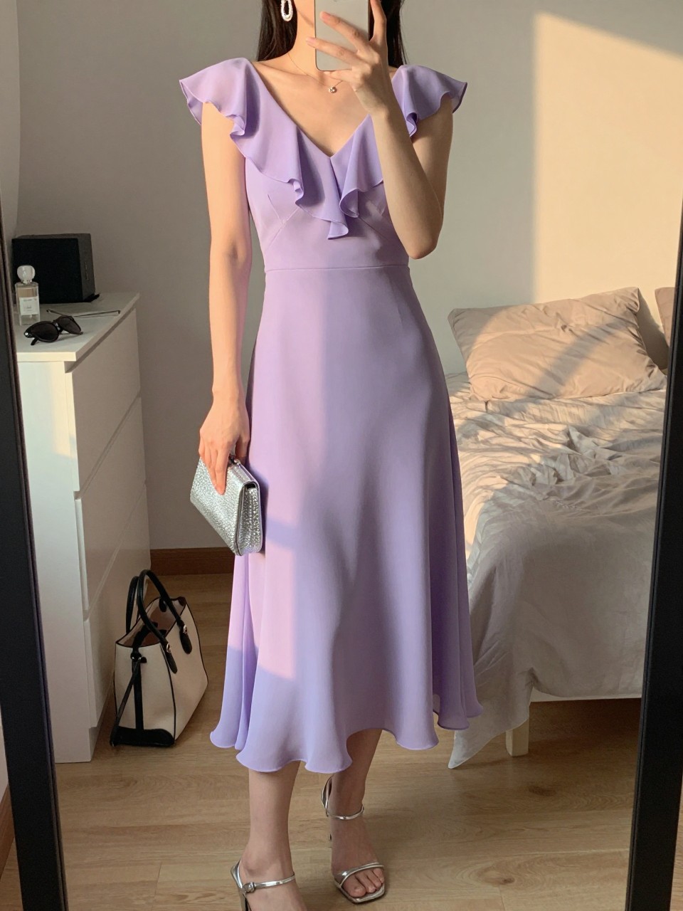 mirror selfie of a woman wearing a lavender chiffon midi dress with soft ruffles, silver drop earrings, silver strappy heels, and a small silver clutch, holding a phone in front of a mirror, cropped at NECK so face doesn't show, with a natural casual selfie pose, in a feminine-themed bedroom, evening golden hour, including small lived-in details like a tote bag on the floor, sunglasses on a dresser, or soft bedding in the background, latest iPhone photo quality.