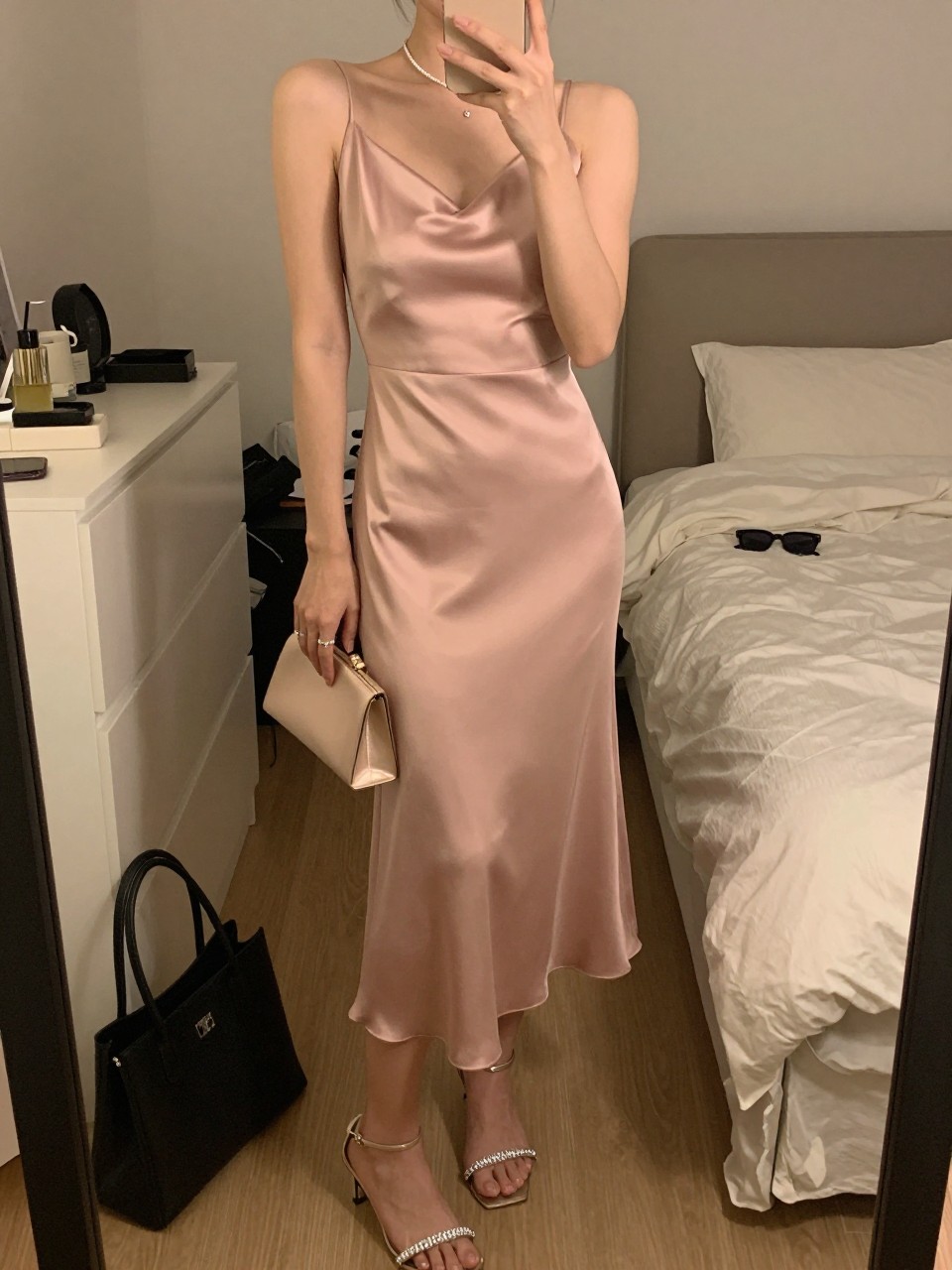 mirror selfie of a woman wearing a blush pink satin midi dress, crystal strap heels, simple crystal stud earrings, and a small blush clutch, holding a phone in front of a mirror, cropped at NECK so face doesn't show, with a natural casual selfie pose, in a feminine-themed bedroom, warm moody lighting, including small lived-in details like a tote bag on the floor, sunglasses on a dresser, or soft bedding in the background, latest iPhone photo quality.