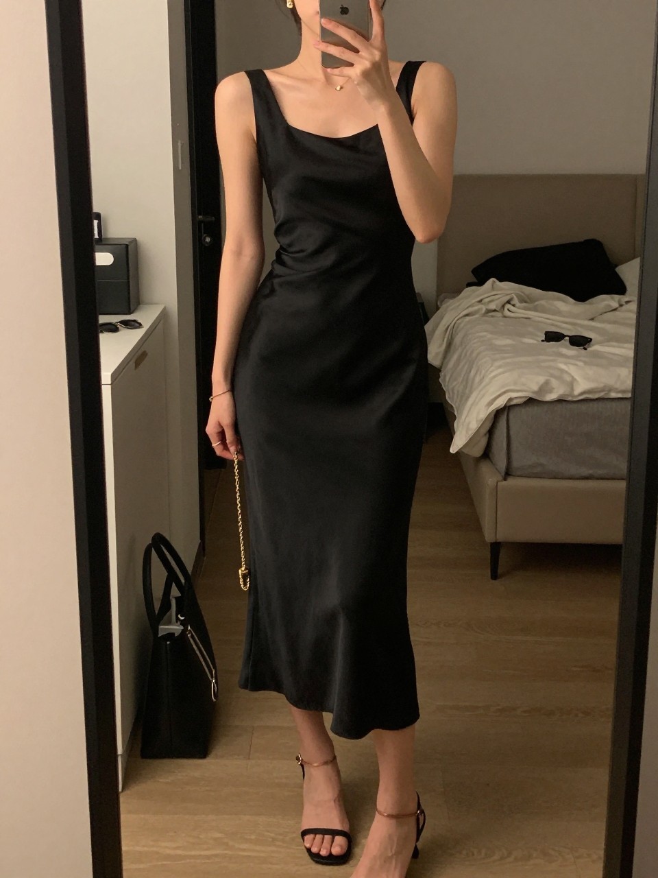 mirror selfie of a woman wearing a black satin midi dress with a clean neckline, black strappy heels, small gold drop earrings, and a gold chain clutch, holding a phone in front of a mirror, cropped at NECK so face doesn't show, with a natural casual selfie pose, in a city apartment, warm moody lighting, including small lived-in details like a tote bag on the floor, sunglasses on a dresser, or soft bedding in the background, latest iPhone photo quality.