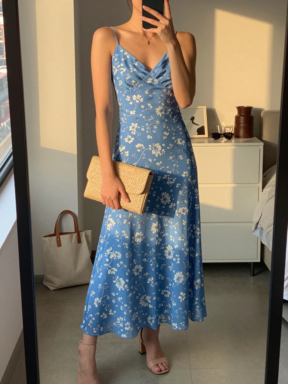 mirror selfie of a woman wearing a blue floral chiffon maxi dress, nude block-heel sandals, a delicate gold pendant necklace, and a woven clutch with a clean shape, holding a phone in front of a mirror, cropped at NECK so face doesn't show, with a natural casual selfie pose, in a city apartment, evening golden hour, including small lived-in details like a tote bag on the floor, sunglasses on a dresser, or soft bedding in the background, latest iPhone photo quality.
