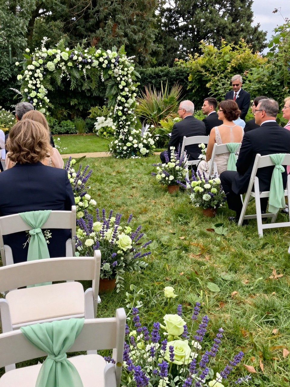 Photo of a late summer garden ceremony aisle with lavender and soft green floral arrangements, cream chairs, and light greenery accents, camera angle: wide ceremony view, setting: outdoor garden ceremony space, lighting: natural daylight, containing subtle real wedding activity such as guests taking seats and a small bouquet resting on a chair, latest iPhone photo quality.