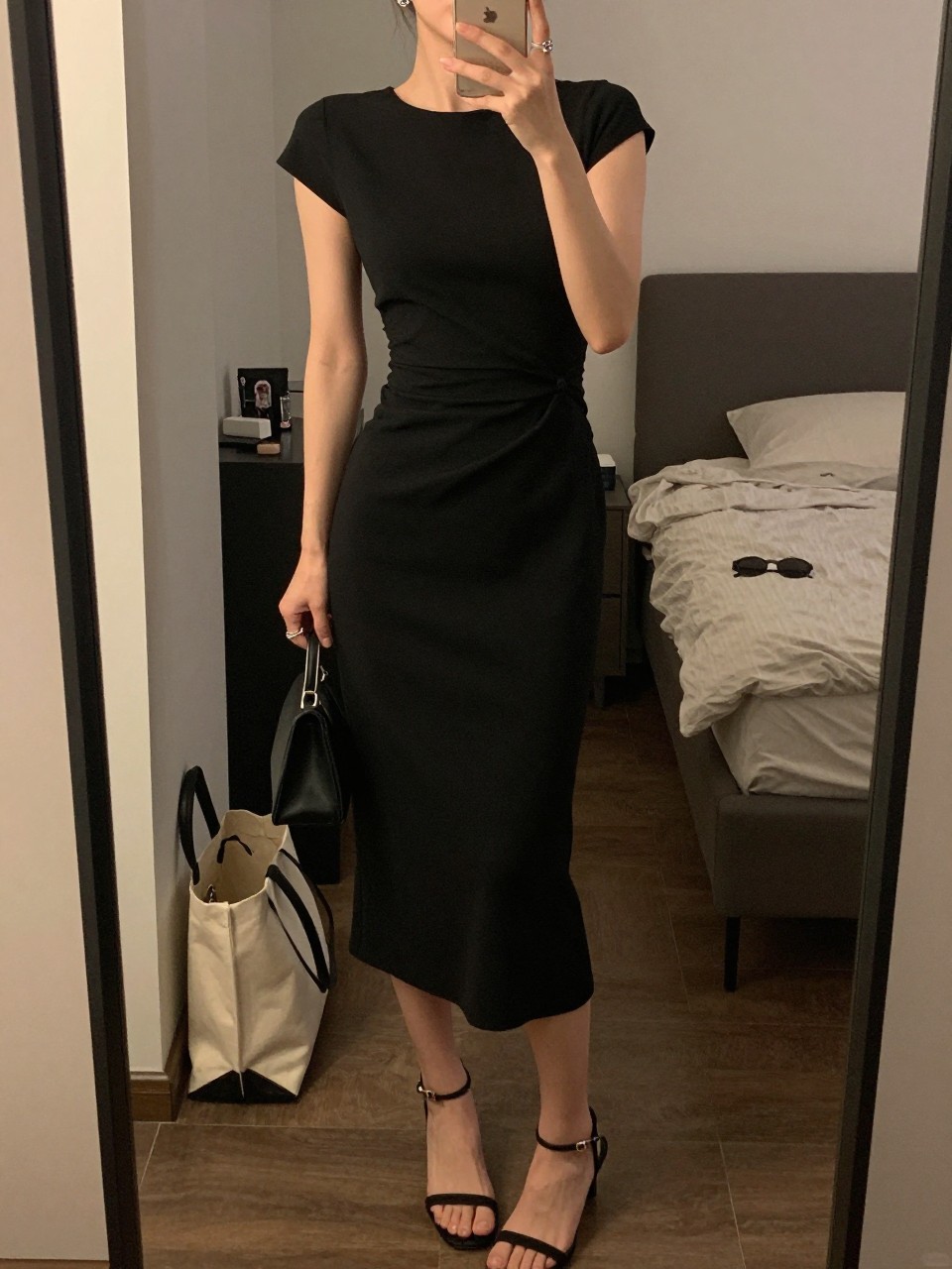 mirror selfie of a woman wearing a black crepe midi dress with a fitted waist, black strappy heels, small crystal stud earrings, and a small black clutch, holding a phone in front of a mirror, cropped at NECK so face doesn't show, with a natural casual selfie pose, in a city apartment, warm moody lighting, including small lived-in details like a tote bag on the floor, sunglasses on a dresser, or soft bedding in the background, latest iPhone photo quality.