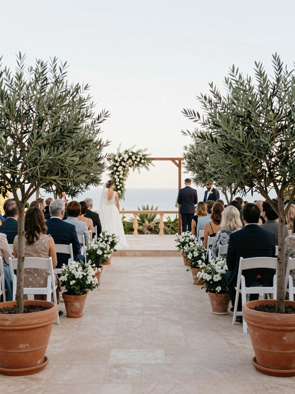 Photo of a ceremony aisle lined with potted olive trees in terracotta planters leading to a simple floral focal point on a Mediterranean terrace, camera angle: wide ceremony view, setting: terrace ceremony aisle, lighting: natural daylight, containing subtle real wedding activity such as the couple walking past at the end of the aisle while guests turn to watch, latest iPhone photo quality.