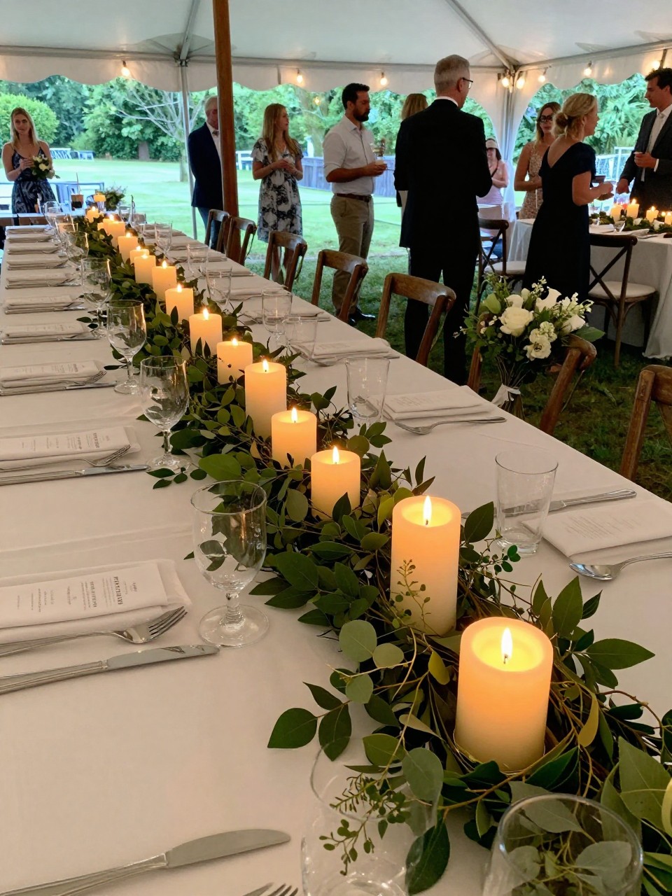 Photo of a summer reception table with a low greenery garland down the center and mixed-height candles glowing among the leaves, camera angle: guest table angle, setting: outdoor tent reception tables, lighting: candlelight glow, containing subtle real wedding activity such as guests chatting and a bouquet resting on a nearby chair, latest iPhone photo quality.