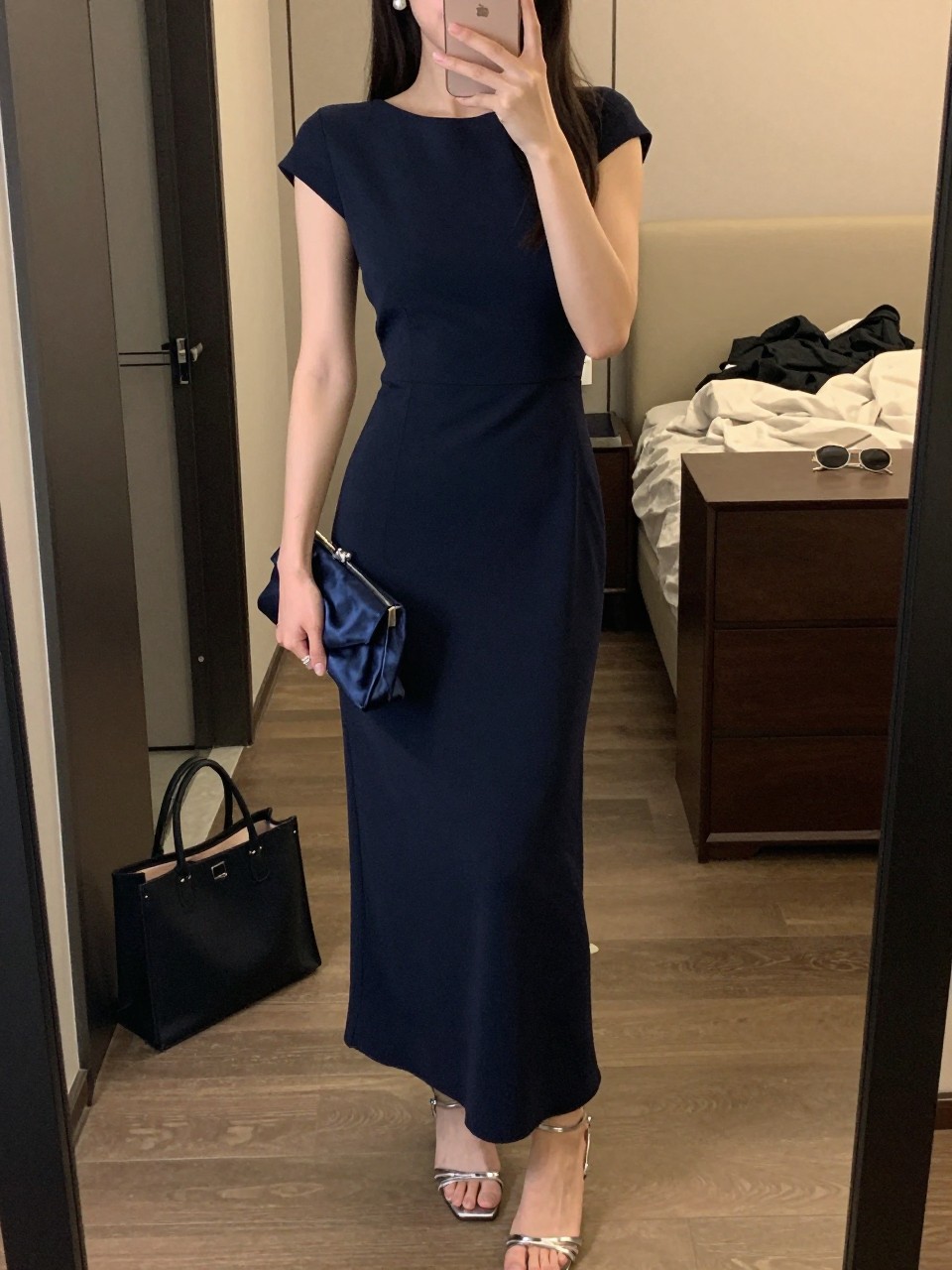 mirror selfie of a woman wearing a navy crepe floor-length column gown, pearl stud earrings, silver strappy heels, and a navy satin clutch, holding a phone in front of a mirror, cropped at NECK so face doesn't show, with a natural casual selfie pose, in a city apartment, warm moody lighting, including small lived-in details like a tote bag on the floor, sunglasses on a dresser, or soft bedding in the background, latest iPhone photo quality.