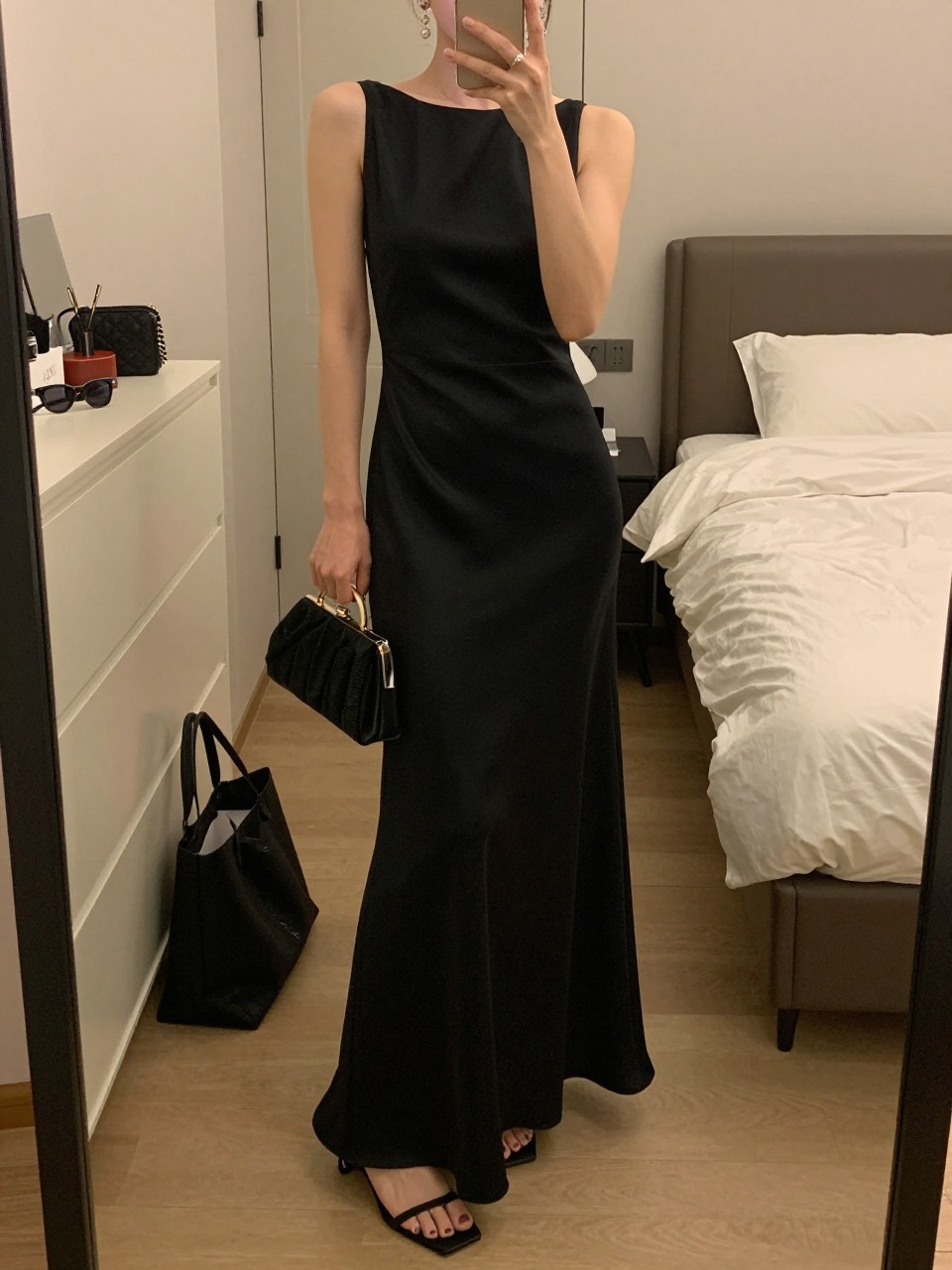 mirror selfie of a woman wearing a floor-length black satin gown with a simple neckline, crystal drop earrings, black strappy heels, and a small structured black clutch, holding a phone in front of a mirror, cropped at NECK so face doesn't show, with a natural casual selfie pose, in a feminine-themed bedroom, warm moody lighting, including small lived-in details like a tote bag on the floor, sunglasses on a dresser, or soft bedding in the background, latest iPhone photo quality.