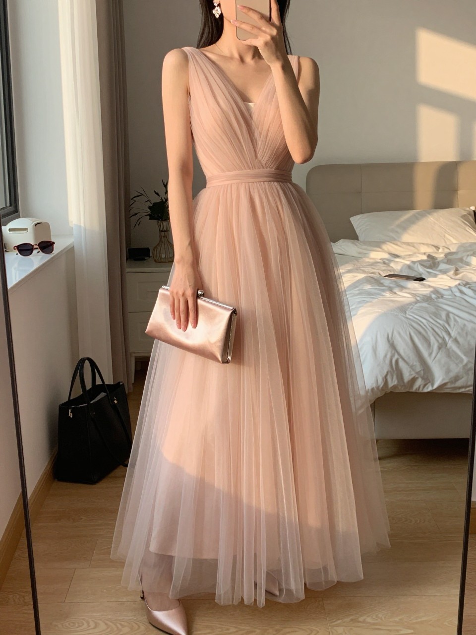 mirror selfie of a woman wearing a blush floor-length tulle gown with a fitted bodice and layered skirt, nude satin heels, delicate drop earrings, and a blush satin clutch, with a rhinestone hair clip visible, holding a phone in front of a mirror, cropped at NECK so face doesn't show, with a natural casual selfie pose, in a feminine-themed bedroom, evening golden hour, including small lived-in details like a tote bag on the floor, sunglasses on a dresser, or soft bedding in the background, latest iPhone photo quality.