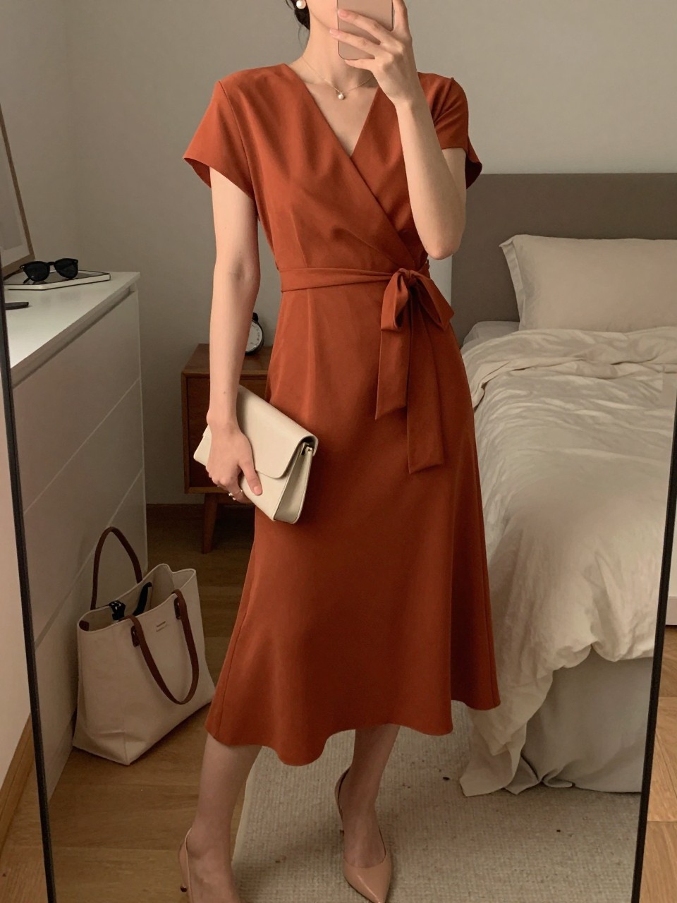 mirror selfie of a woman wearing a terracotta crepe wrap midi dress with a tied waist, nude slingback heels, small pearl stud earrings, and a small cream clutch, holding a phone in front of a mirror, cropped at NECK so face doesn't show, with a natural casual selfie pose, in a feminine-themed bedroom, warm moody lighting, including small lived-in details like a tote bag on the floor, sunglasses on a dresser, or soft bedding in the background, latest iPhone photo quality.