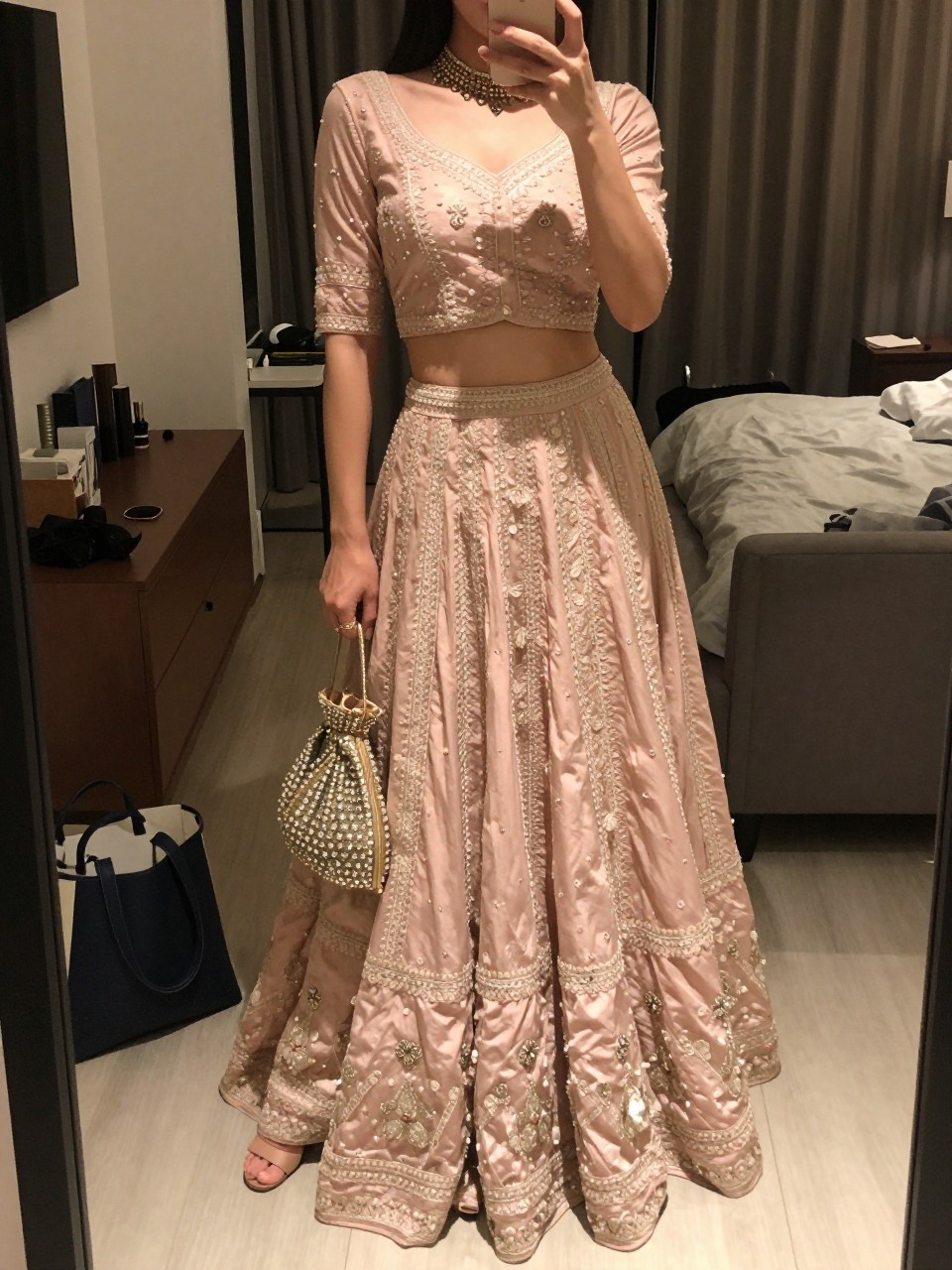 mirror selfie of a woman wearing a blush lehenga with light embroidery, a fitted matching choli, a stone choker necklace, nude block heels, and an embellished potli bag, holding a phone in front of a mirror, cropped at NECK so face doesn't show, with a natural casual selfie pose, in a city apartment, warm moody lighting, including small lived-in details like a tote bag on the floor, sunglasses on a dresser, or soft bedding in the background, latest iPhone photo quality.