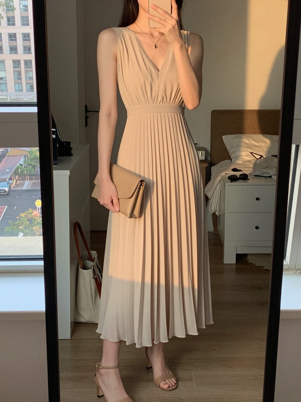 mirror selfie of a woman wearing a beige pleated chiffon midi dress, a delicate gold pendant necklace, tan heeled sandals, and a small tan clutch, holding a phone in front of a mirror, cropped at NECK so face doesn't show, with a natural casual selfie pose, in a city apartment, evening golden hour, including small lived-in details like a tote bag on the floor, sunglasses on a dresser, or soft bedding in the background, latest iPhone photo quality.