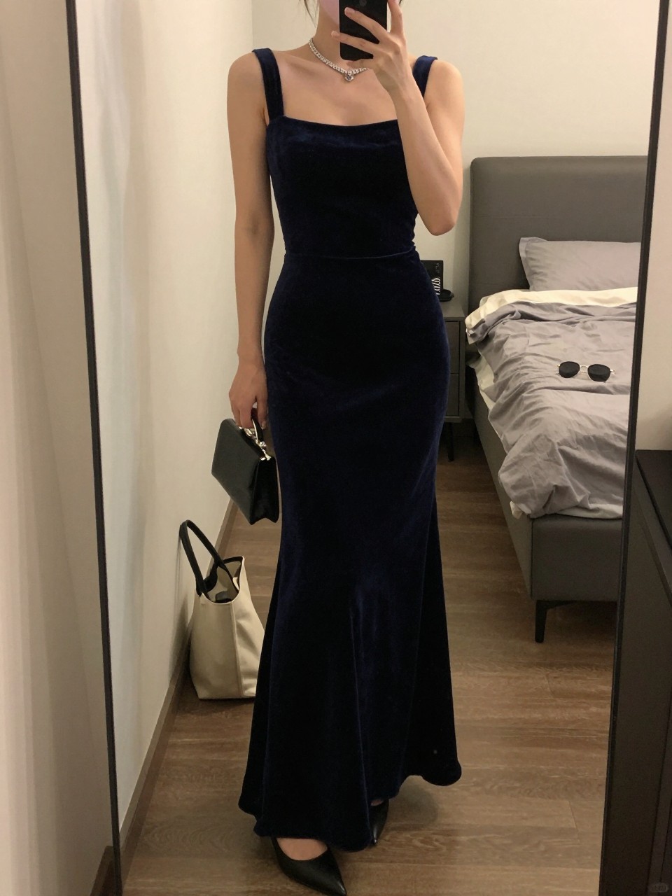 mirror selfie of a woman wearing a midnight blue velvet floor-length gown with a fitted shape and a slit, a diamond tennis necklace, black pointed-toe heels, and a small black box clutch, holding a phone in front of a mirror, cropped at NECK so face doesn't show, with a natural casual selfie pose, in a city apartment, warm moody lighting, including small lived-in details like a tote bag on the floor, sunglasses on a dresser, or soft bedding in the background, latest iPhone photo quality.