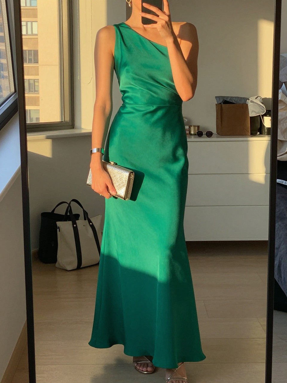 mirror selfie of a woman wearing an emerald one-shoulder floor-length gown in satin or crepe, metallic strappy heels, a slim cuff bracelet, and a metallic clutch, holding a phone in front of a mirror, cropped at NECK so face doesn't show, with a natural casual selfie pose, in a city apartment, evening golden hour, including small lived-in details like a tote bag on the floor, sunglasses on a dresser, or soft bedding in the background, latest iPhone photo quality.