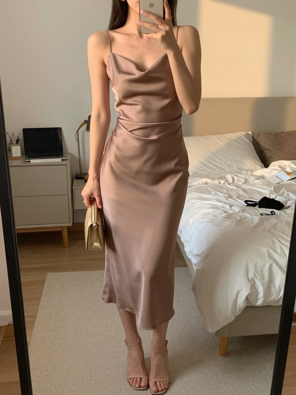 mirror selfie of a woman wearing a mocha satin midi dress with a draped skirt, nude strappy heels, small gold stud earrings, and a small gold clutch, holding a phone in front of a mirror, cropped at NECK so face doesn't show, with a natural casual selfie pose, in a feminine-themed bedroom, evening golden hour, including small lived-in details, latest iPhone photo quality.